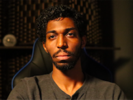 Andrew looks at the camera while wearing a black T-shirt and sitting in a black gaming chair with blue accents.