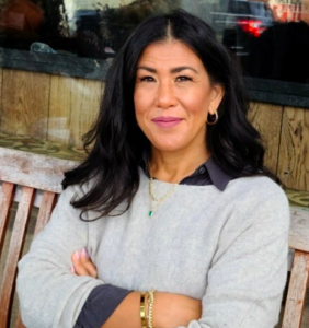 Kilmeny crosses her arms and smiles at the camera, wearing a gray sweater over a navy button-up with gold jewelry.