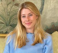 Isabel softly smiles while wearing a light blue top in front of a painting of green foliage. 