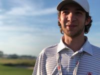 Desi grins while on a golf green, wearing a white-striped polo and hat.