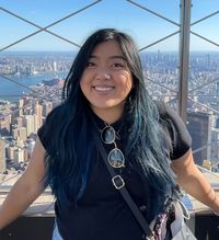 Halle grins at the camera, sunglasses clipped to her shirt and blue highlights in her hair, while standing in front of the New York City skyline.