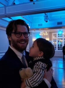 A white mane with mid-length brown hair and a beard, holds a child and grins widely at the camera.