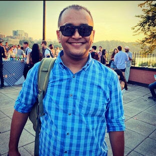 A man wearing a blue plaid shirt and dark sunglasses smiles in front of a beautiful scene on a riverside.