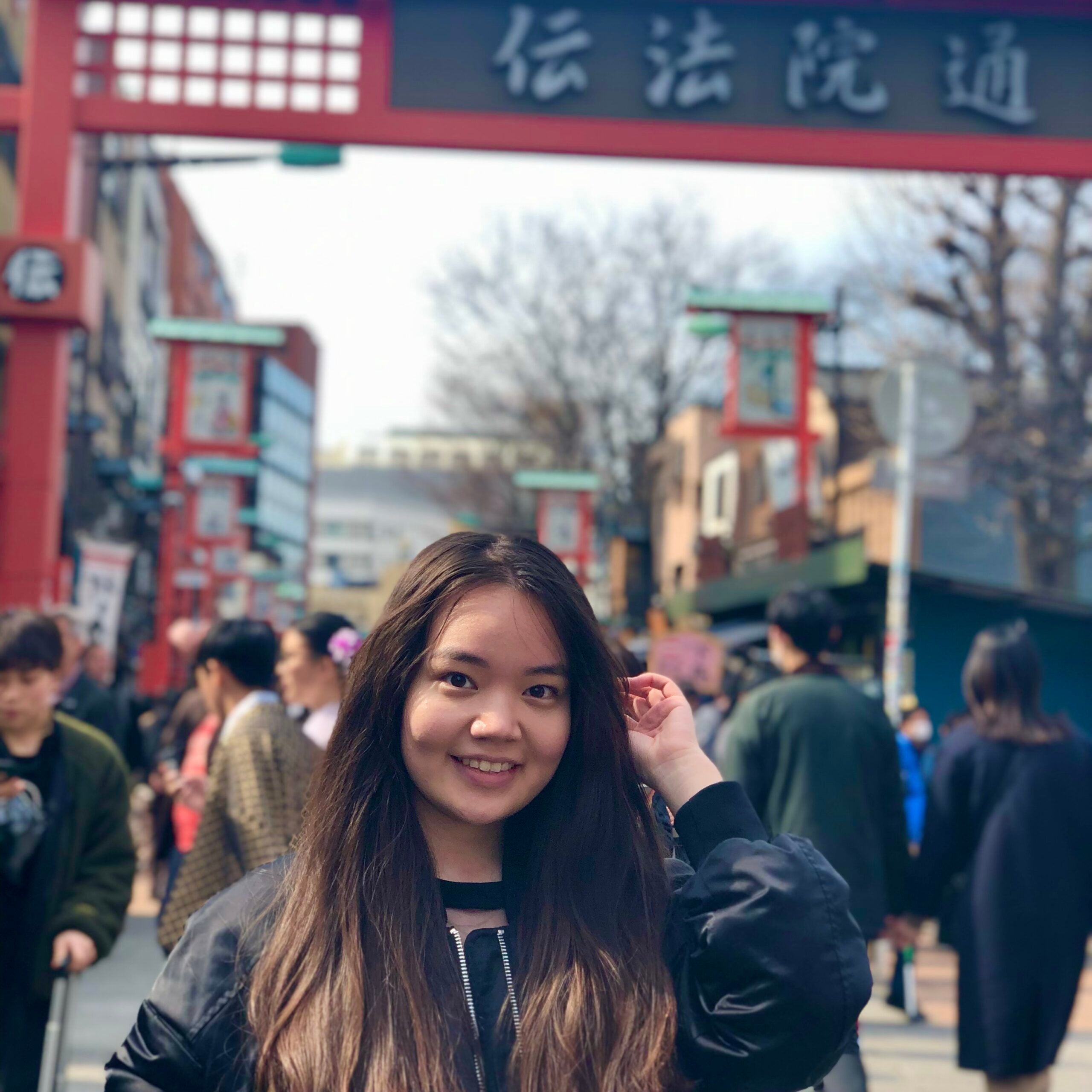 Astiya smiles at the camera and plays with her hair in front of a red gate. Other city-goers are visible behind her.