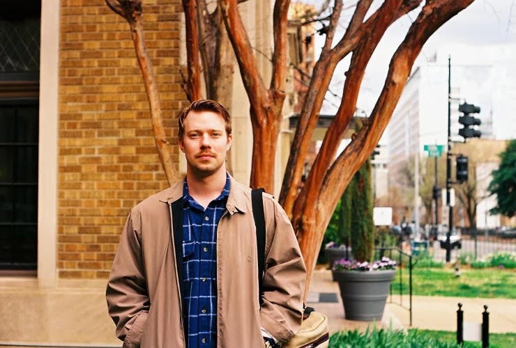 Hayden looks at the camera from across the street. He is dressed in a blue collared shirt and a tan jacket and he has his hands in his pockets.