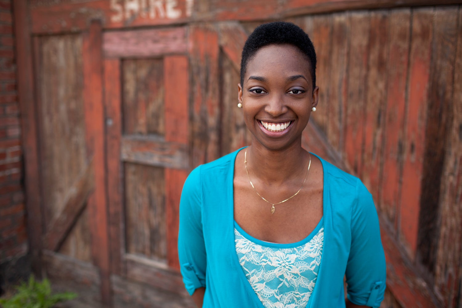 The image depicts a smiling brown-skinned woman with short dark hair wearing a teal and cream blouse who is standing in front of a red, distressed wood backdrop.
