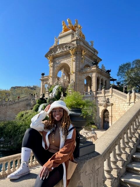 Kyra sits on a concrete handrail while smiling. Behind her, an ornate concrete and brick structure is visible against a blue sky.