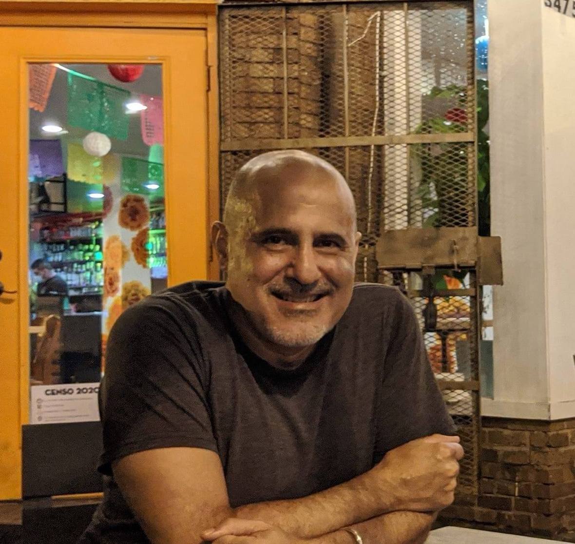 A headshot of a brown-skinned bald man sitting in front of a bodega with his arms crossed and resting on a table. He is wearing a brown t-shirt and smiling at the camera.