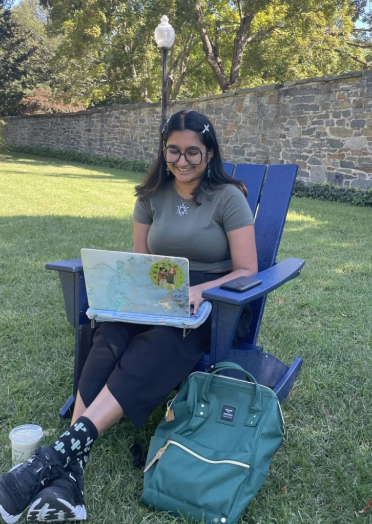 Kavita smiles down at her open laptop, perched on her knees, while she sits in a blue rocking chair in Healy Lawn. 