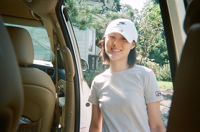 June wears a white baseball hat and smiles at the camera, looking into the back of a vehicle.