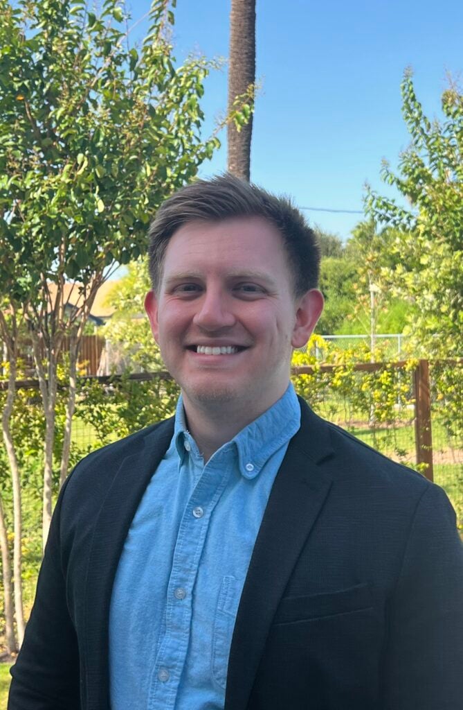 Andrew smiles at the camera while wearing a blue button up shirt and blazer, posing in front of a sunny ivy-covered fence