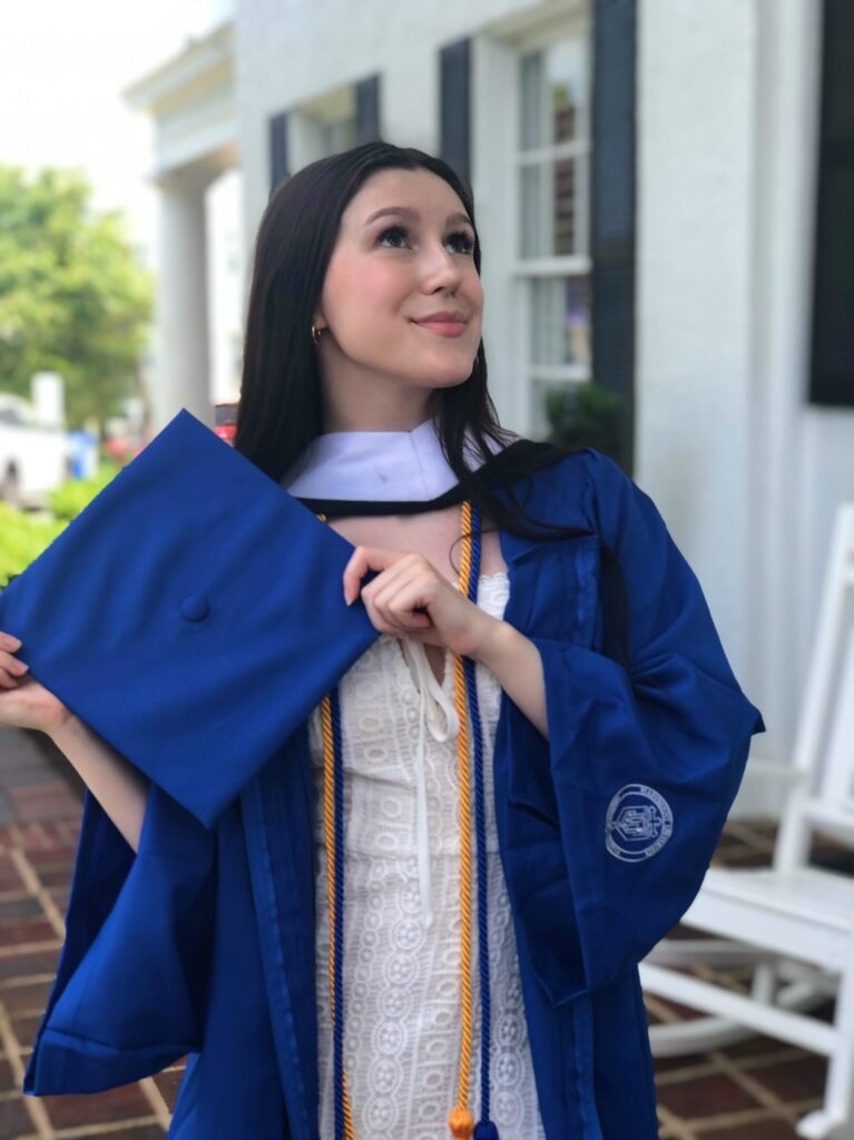 Bella smiles while looking up, proudly holding her brilliant blue graduation cap in front of her. 