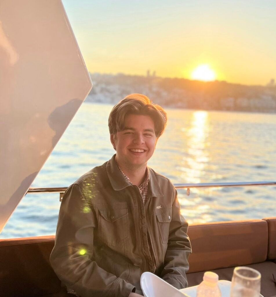 Sam grins at the camera, posed in front of a waterfront view with the sunset in the background.