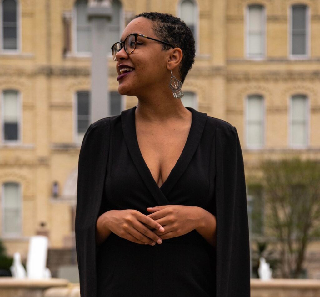 Jamira looks to her right with a grin on her face while wearing a black business outfit. 