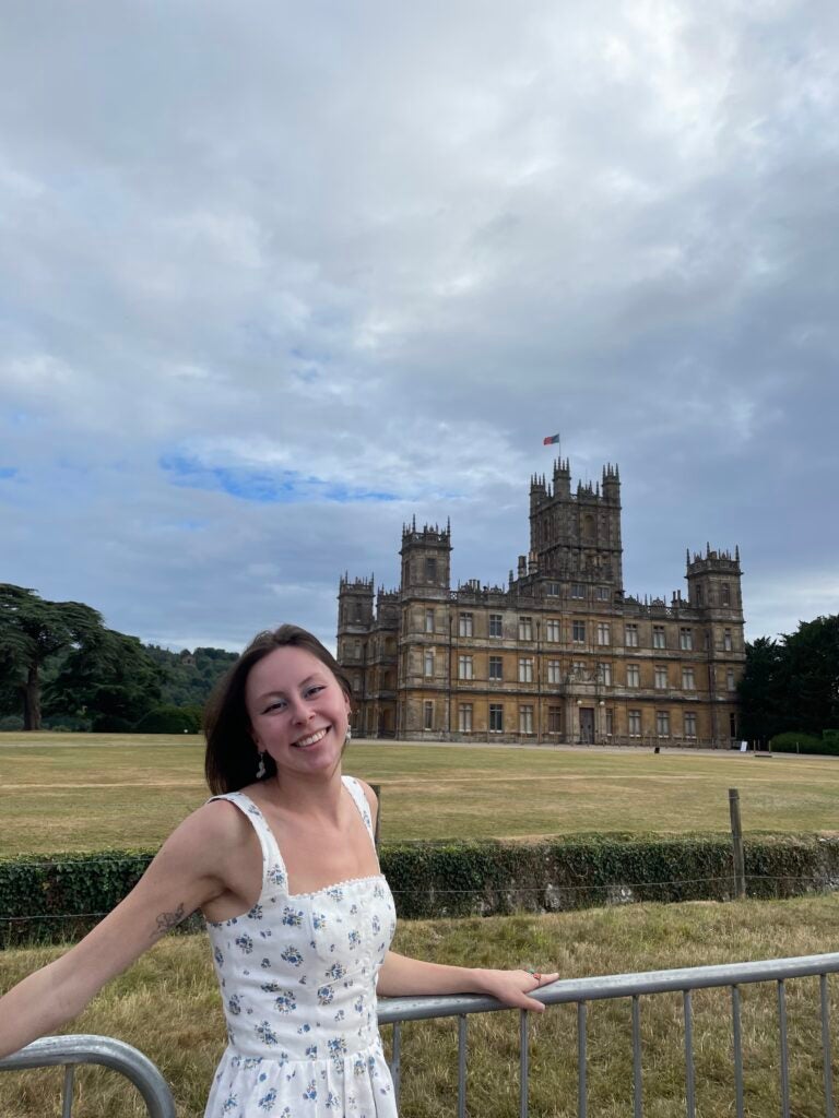 Olivia tips her head back and smiles, wearing a white and blue dress, while in front of a castle on an overcast day.