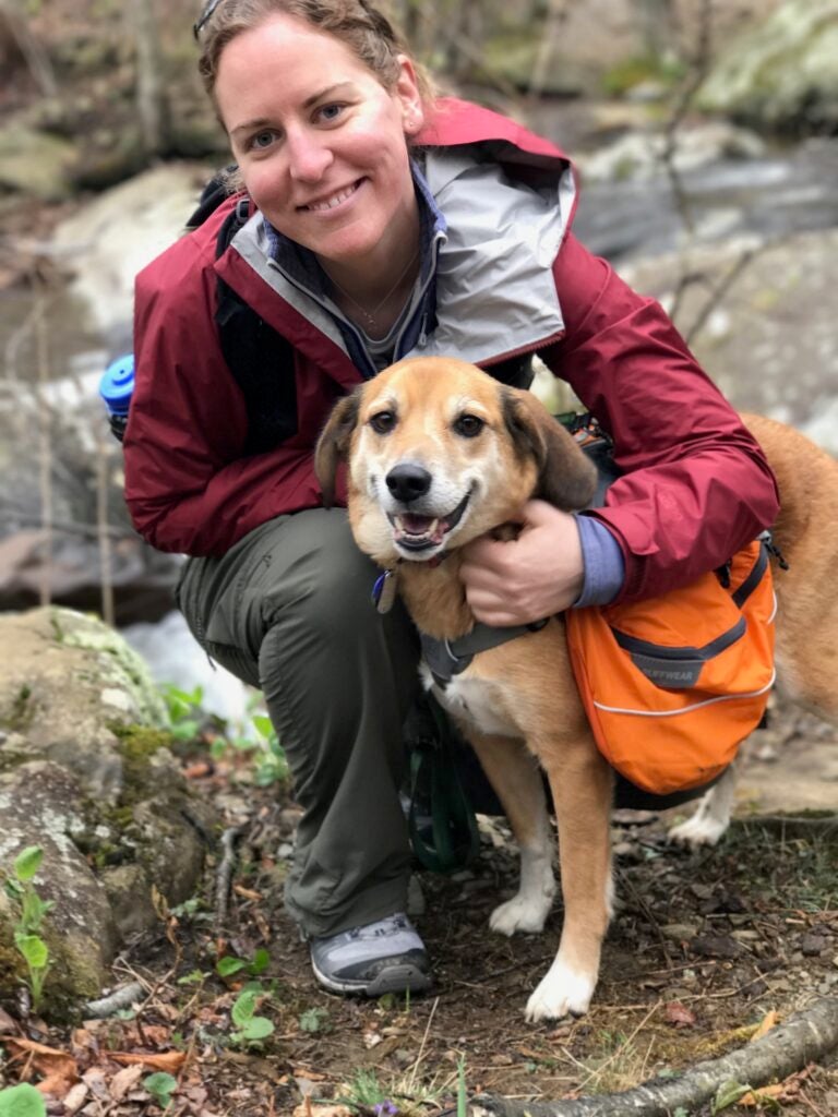 Meghan smiles and looks up at the camera while posing with a medium-sized brown dog. 