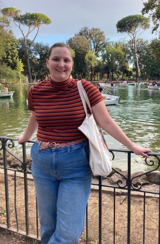 Rebecca smiles while leaning against an iron fence in front of a pond in which others are paddling rowboats.