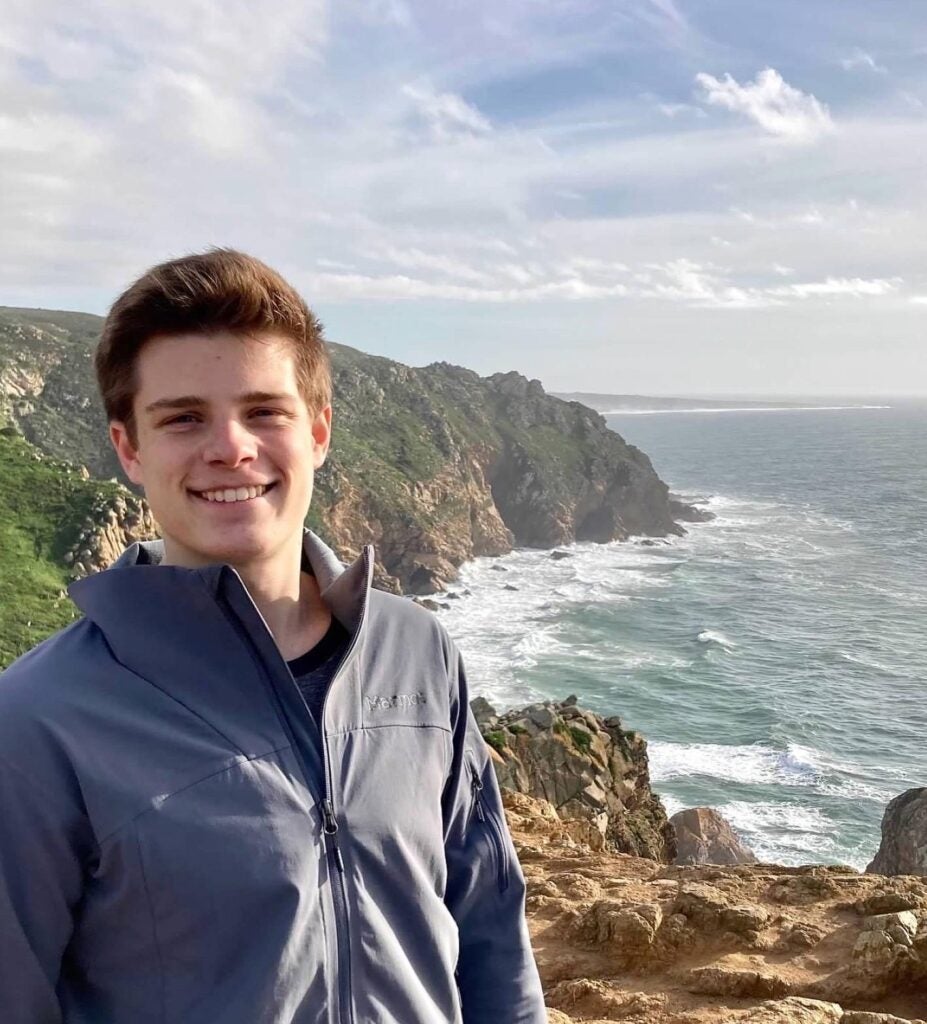 Chase smiles at the camera. Behind him, a rocky coastline and calm seas are visible. 