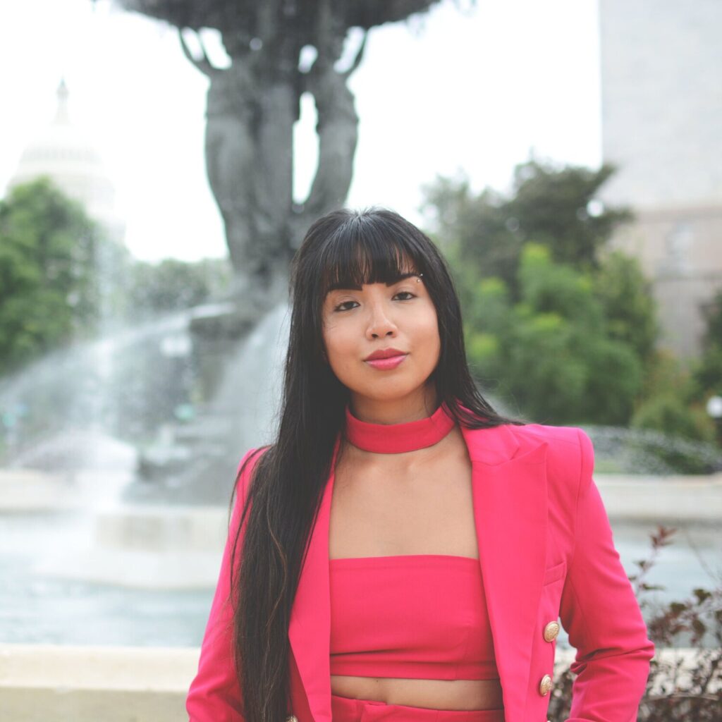 Mekita looks directly at the camera while wearing a bright pink blazer and standing before a fountain.