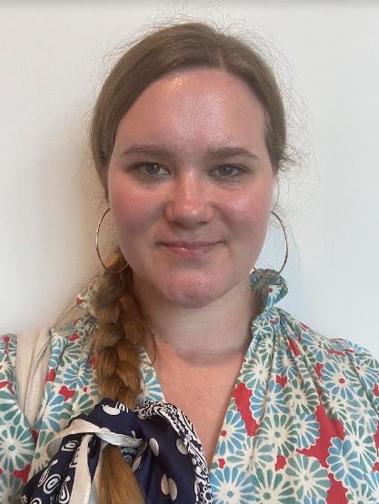 Ellie stands against a white background, wearing a blue and red floral dress, smiling at the camera. 