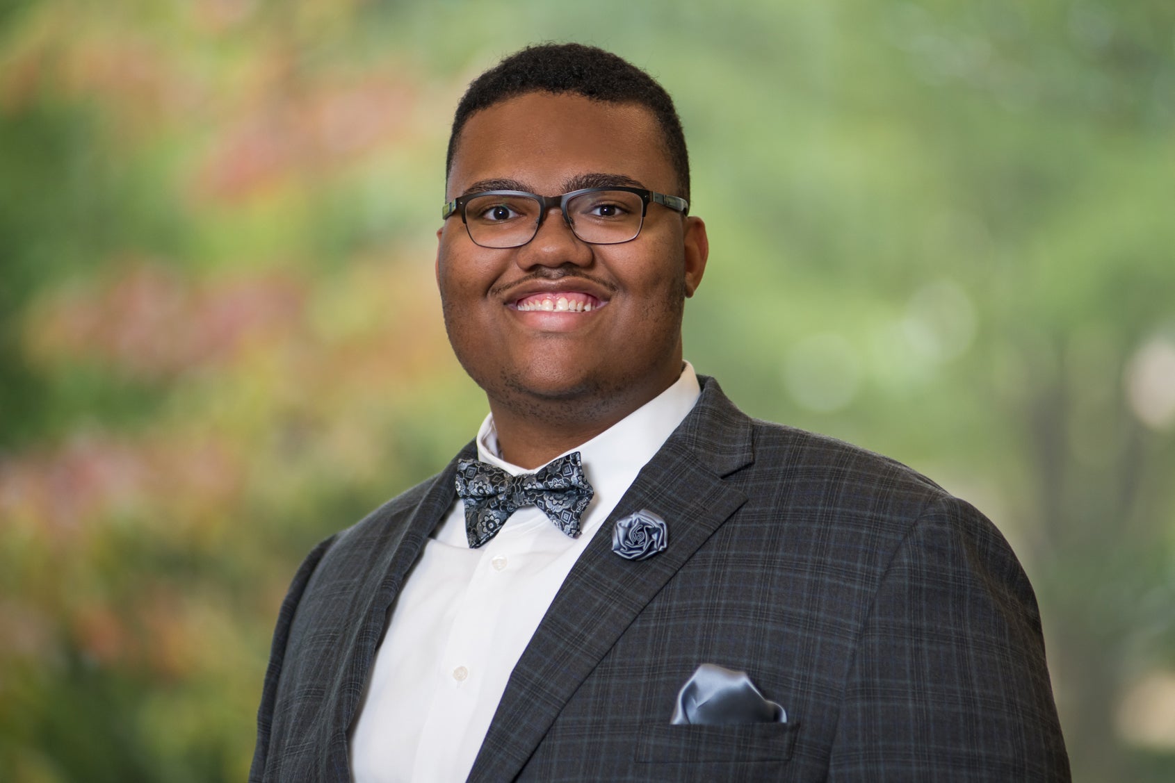 Isaiah stands in a checkered jacket and bowtie, smiling warmly at the camera in front of a green background. 