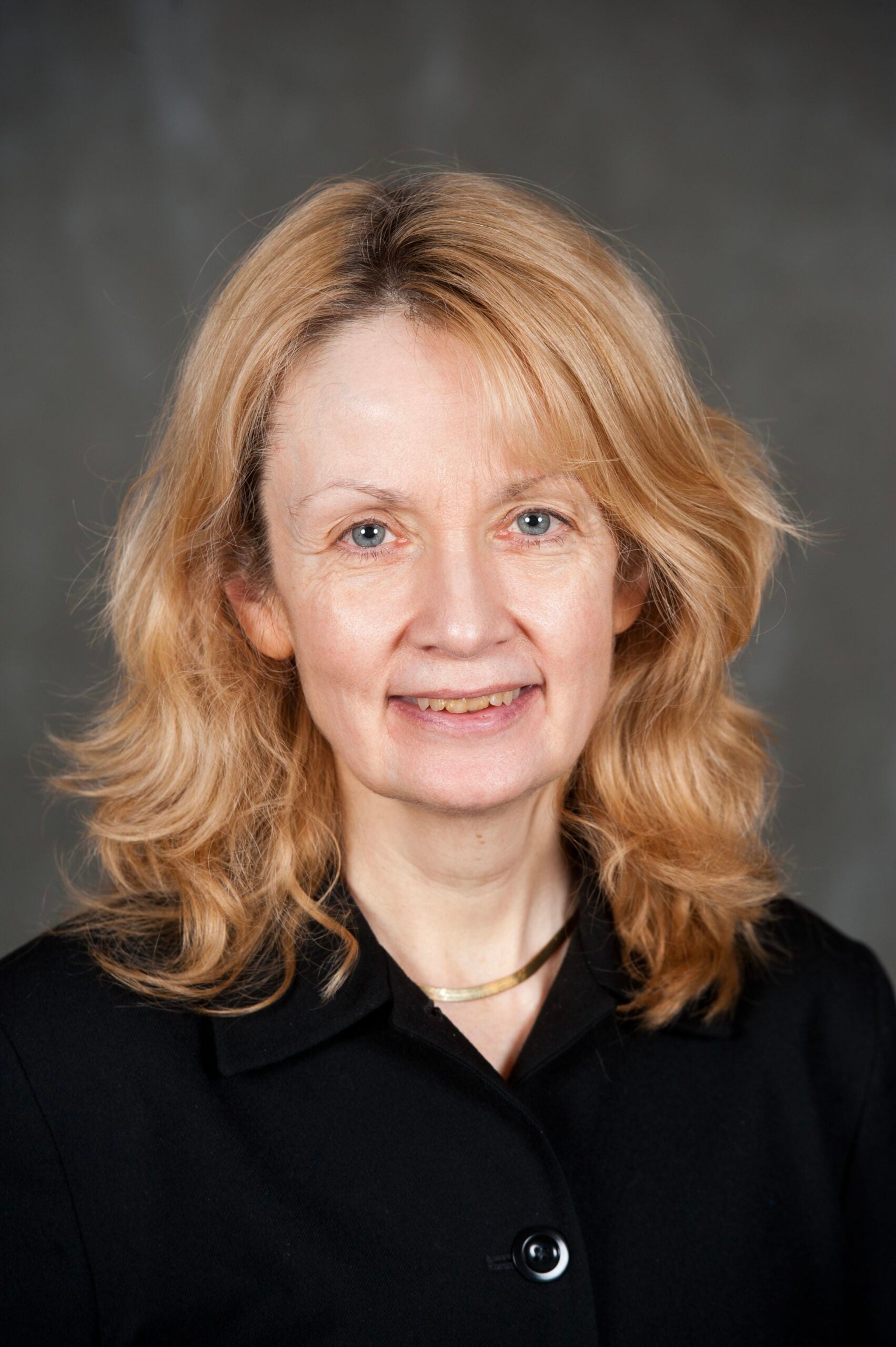 The woman in photo is Caucasian with blue eyes and blond hair. She wears a black suit and gold necklace and gentle smile.
