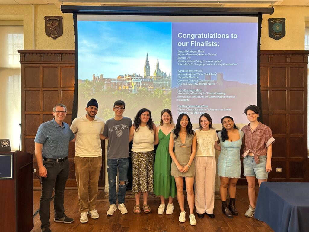 Dr. Phil Sandick, Kirit Minhas, Clayton Kincade, Cassandra Leahy, Genevieve Jobson, Josephine Wu, Maya Kominsky, Alison Karki, and Ollie Shirley smile for a picture beneath the names of our finalists projected on a large screen.