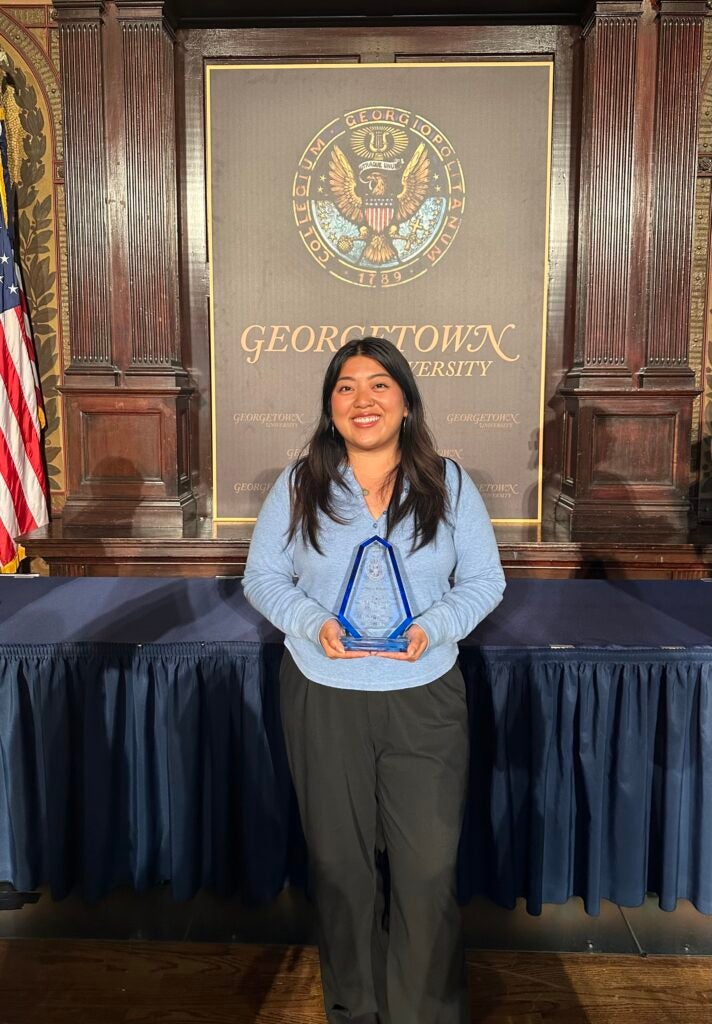 Halle Trang smiles in front of a wooden Georgetown University Seal holding her Graduate Student Award. 