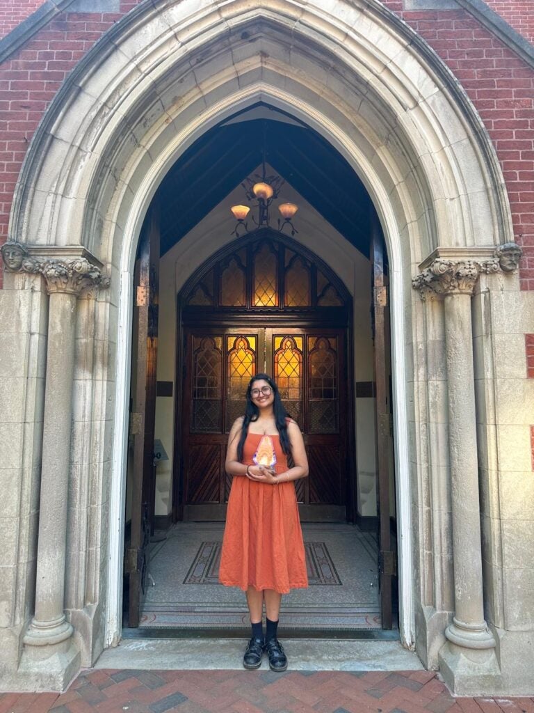 Kavita Premkumar stands in a burnt orange dress, holding her award beneath a vaulted doorway.