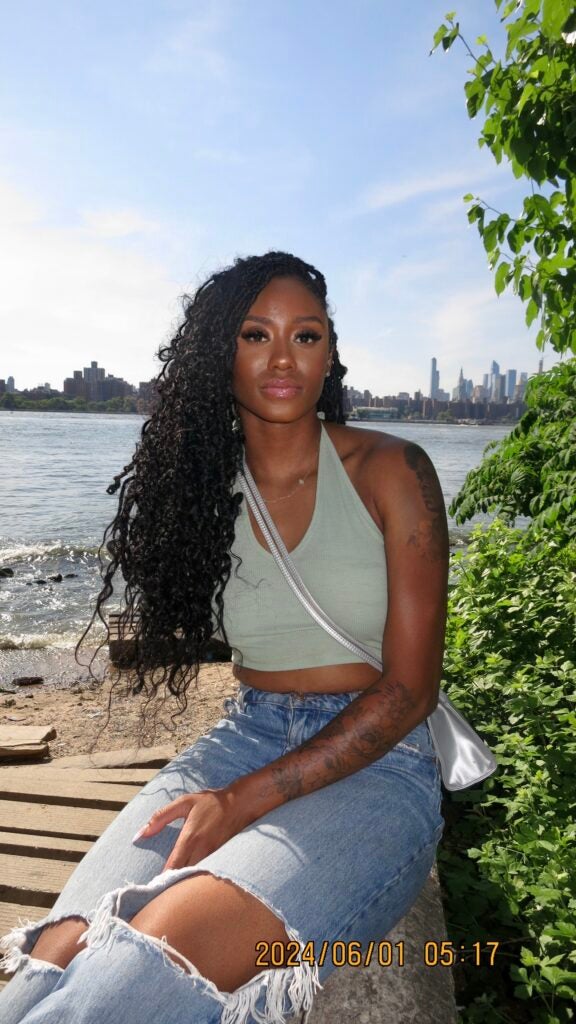Laurah sits on a pier in jeans and a green top, Manhattan and the Hudson River behind her, smiling at the camera. 