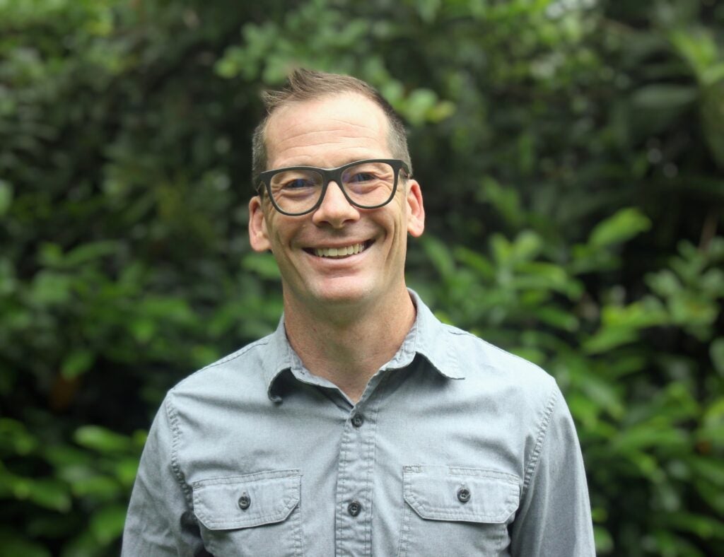 Nathan Hensley smiles in a grey camp shirt and dark wooden glasses, standing in front of a grove of pine trees. 