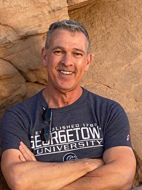 Professor Ortiz stands against a yellow-stone wall, wearing a Georgetown University t-shirt, crossing his arms, and smiling wide at the camera. 
