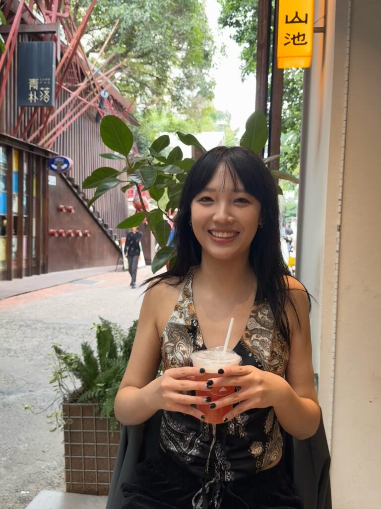Liqian stands on a street in a patterned top holding some a cold beverage, smiling at the camera. 
