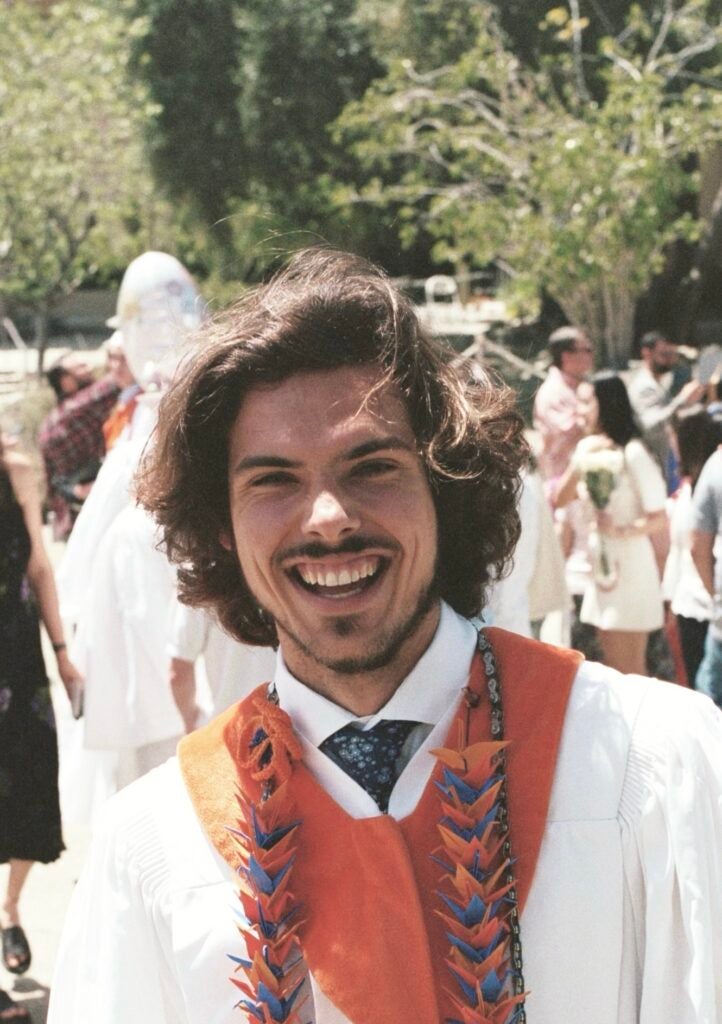 Ryan, wearing a white commencement robe and orange hood, smiles widely at the camera on graduation day. 