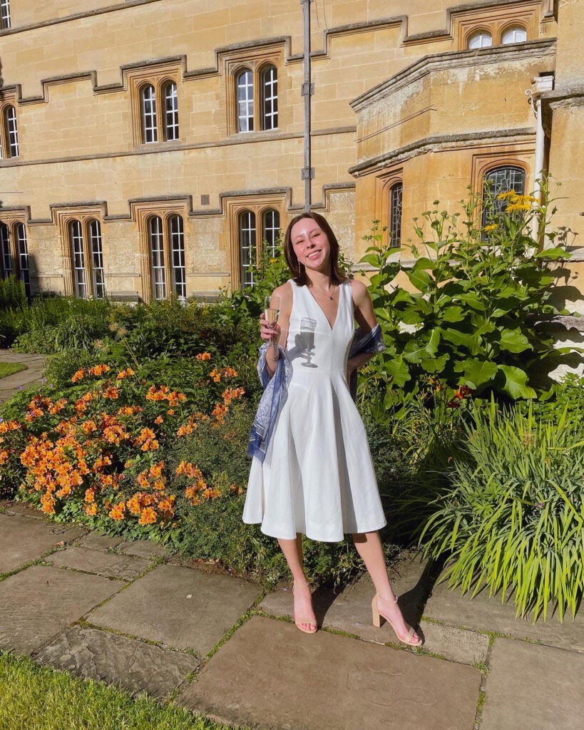 Olivia smiles in a white dress in front of a stone building and courtyard. 
