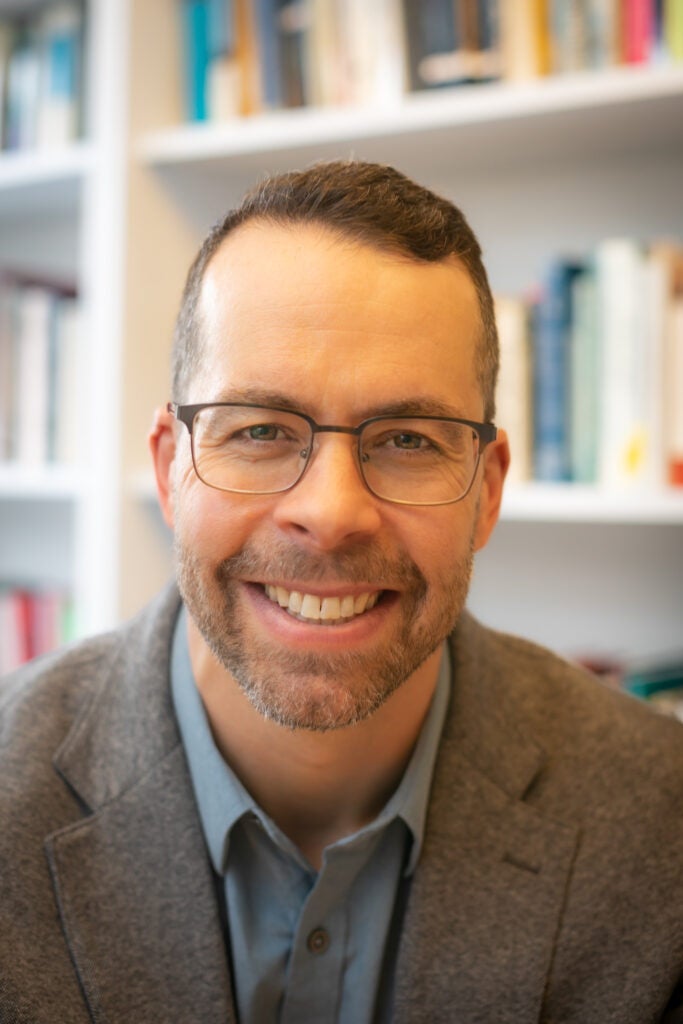 Daniel Shore smiles at the viewer while seated before a white bookcase; he wears a tweed jacket over a grey, collared shirt.