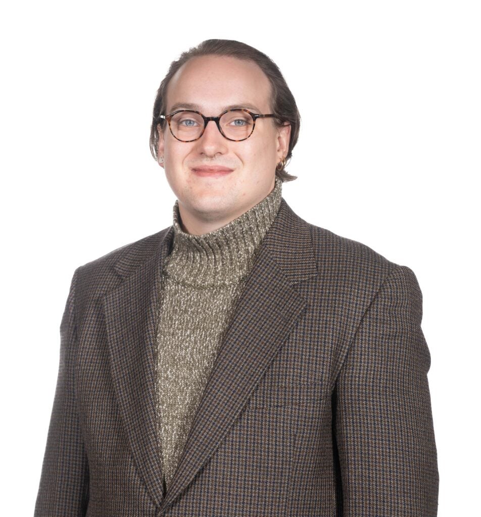 Chris Doyle, a white masculine person wearing tortoise shell glasses wears a checked sport coat over a green turtleneck and smiles softly at the viewer