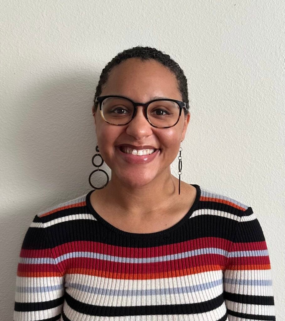 Jamira Richardson, a Black woman with short, curly hair and black, circular, dangly earrings smiles brightly at the camera against a white background. She wears a colorful striped shirt and tortoise shell glasses.
