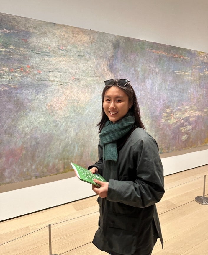 June Yoon smiles at the camera, standing in front of an impressionist painting, wearing a chore coat and green scarf.