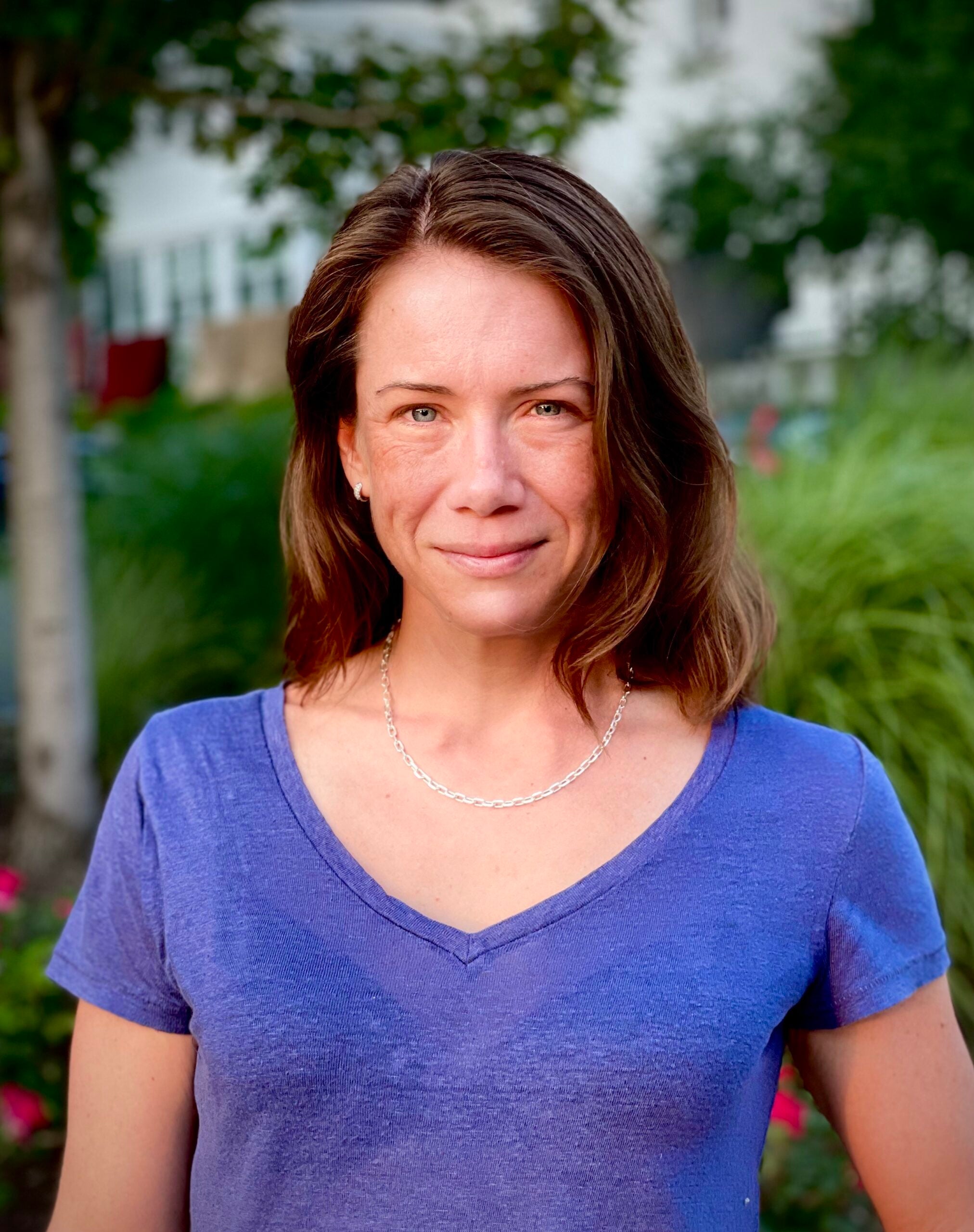 A smiling white woman with brown hair in a blue tee shirt standing in front of several lush green plants.