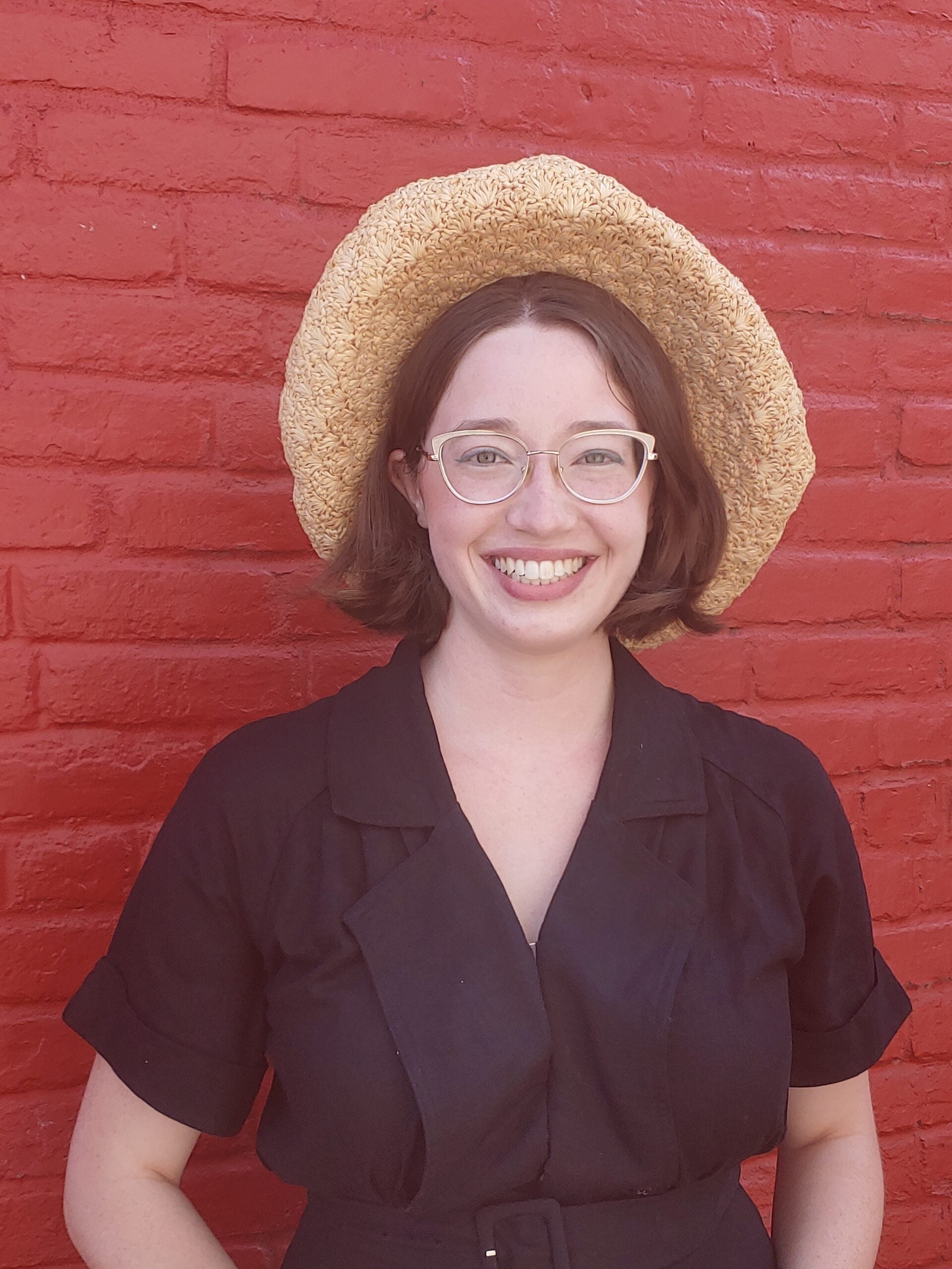 A young woman in glasses and a brunette bob smiles in a black dress and sun hat