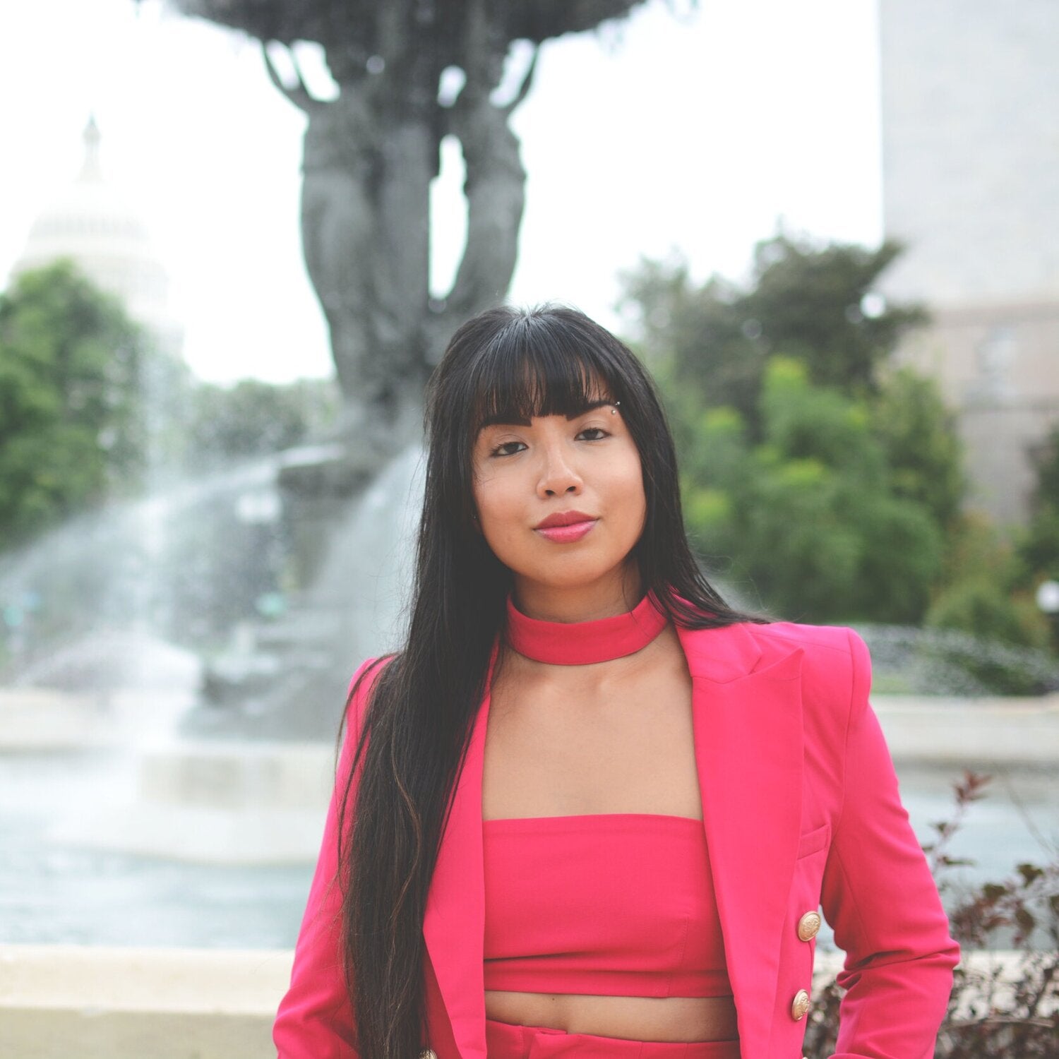 Mekita stands in front of a large fountain, wearing a pink blazer and top, smiling at the viewer. 