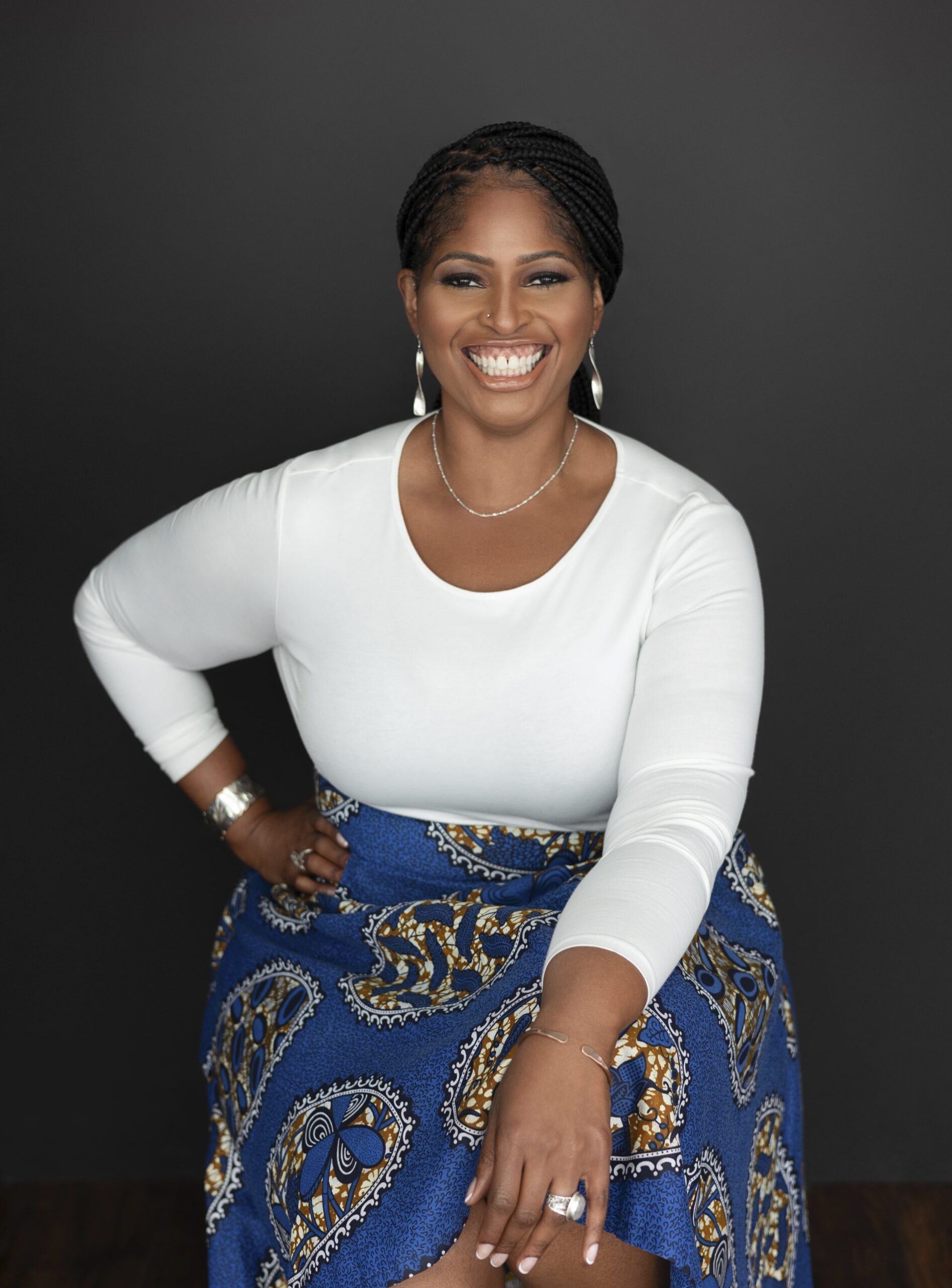 Mecca Jamilah Sullivan wears a white shirt and blue African print skirt against a dark background.