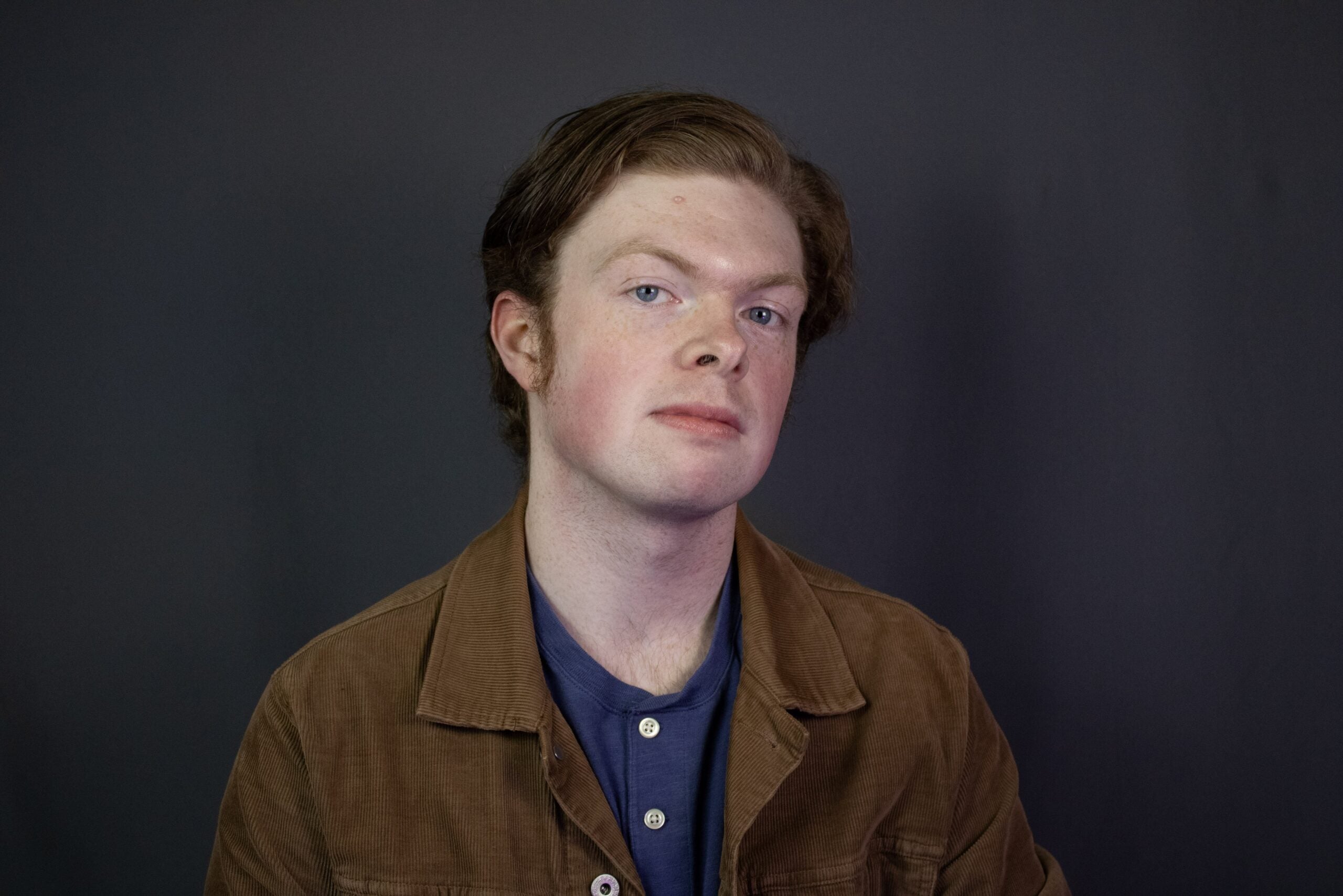 George looks forward, chin tilted up in a brown chore coat and a blue buttoned shirt.