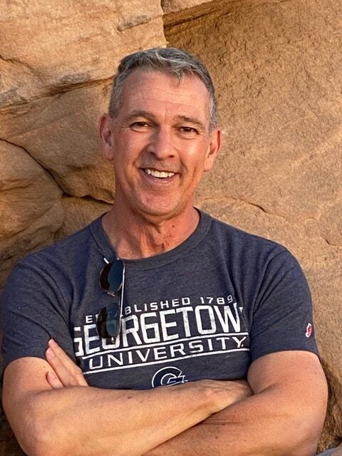Professor Ortiz stands against a yellow-stone wall, wearing a Georgetown University t-shirt, crossing his arms, and smiling wide at the camera.