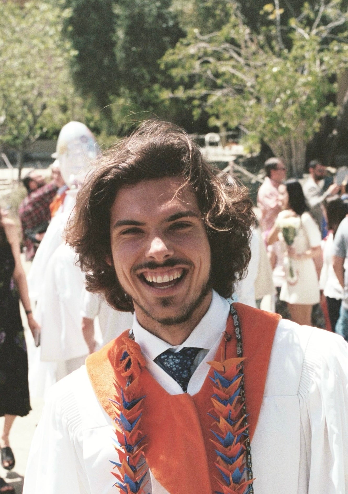 Ryan wearing a white graduation robe and lei smiles broadly at the viewer. 