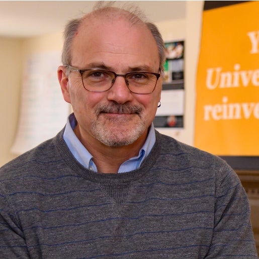 Randall Bass smiles softly at the viewer, wearing a soft gray pullover sweater over a blue collared shirt.