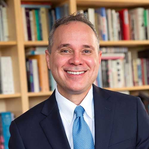 Eddie Maloney stands in front of a bookcase, wearing a sharp, blue suit, white shirt, and blue tie.
