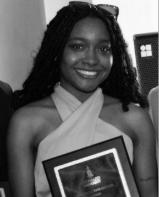 Bria Charlton, wearing a simple light dress holds an award from Howard University and smiles at the camera