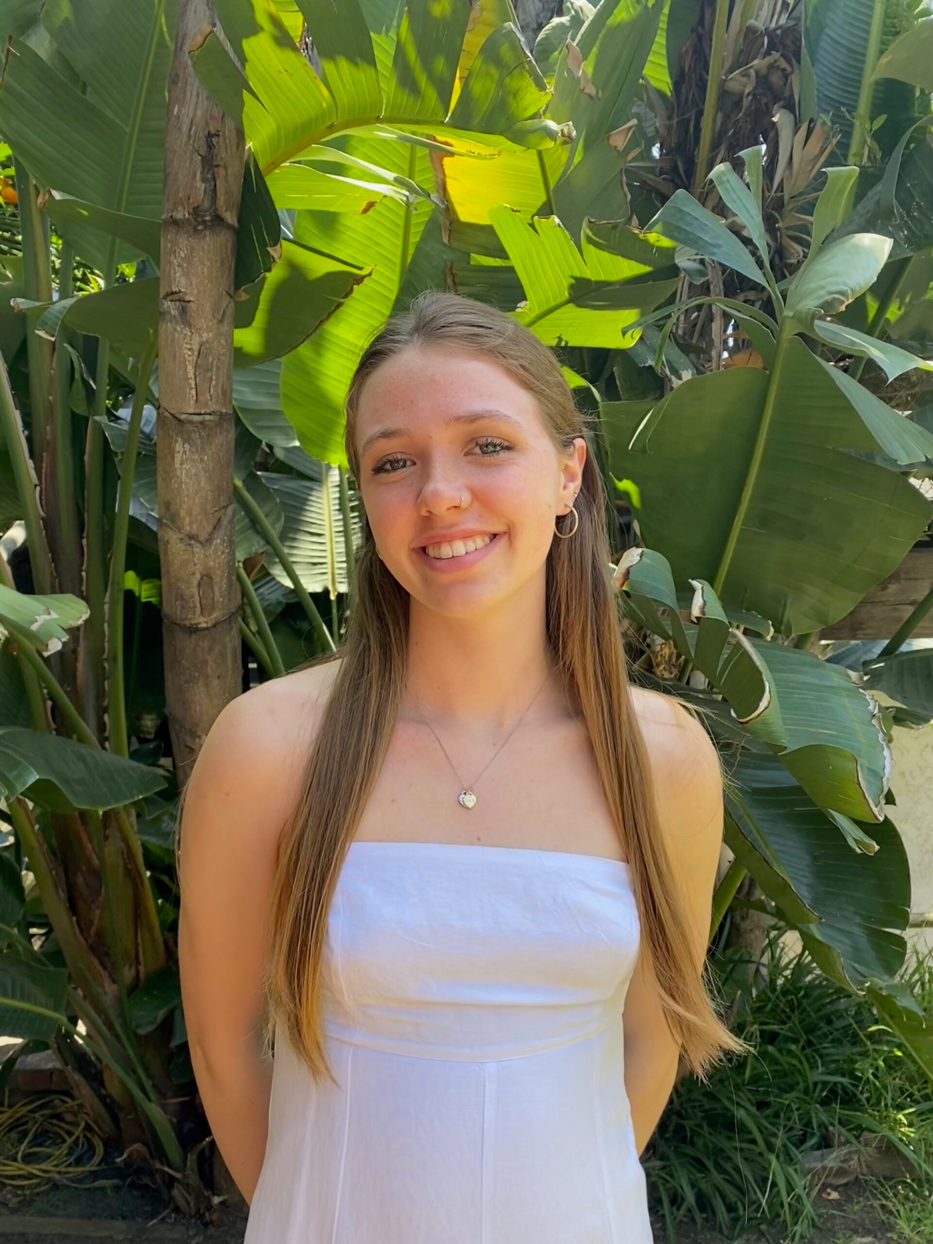 Grace Ringuette wearing  white dress wither her hair down over her shoulders, smiles against a leafy backdrop.